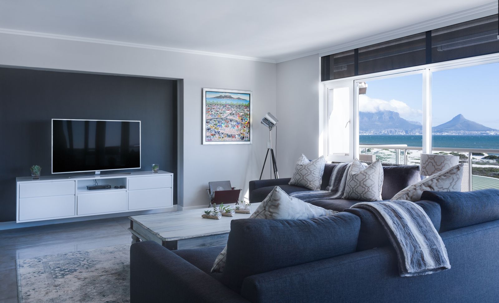 Soggiorno luminoso con divano blu, televisore e grandi vetrate che si aprono su una vista di mare e montagne