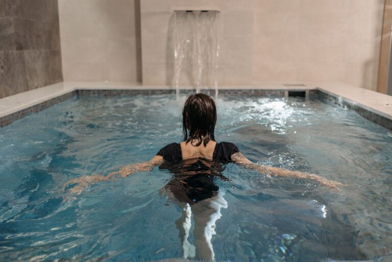 Donna di spalle nella piscina interna di un centro benessere, con una cascata d'acqua sulla parete di pietra
