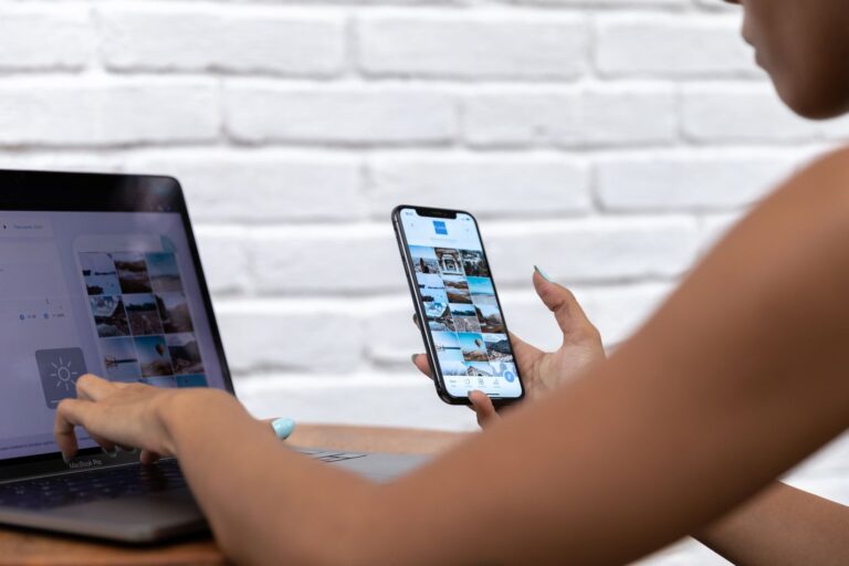 Persona che lavora al portatile e guarda sullo smartphone la stessa galleria di foto, davanti a un muro di mattoni bianchi