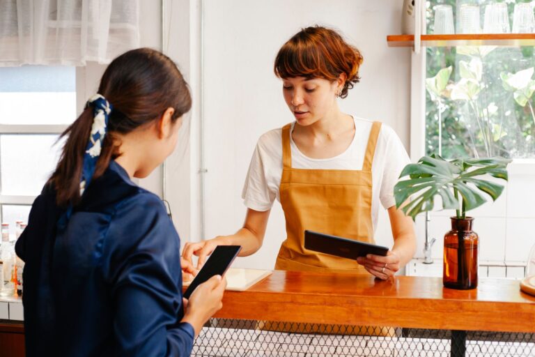 Titolare di un caffè con un tablet al bancone mentre una cliente compila un modulo sullo smartphone