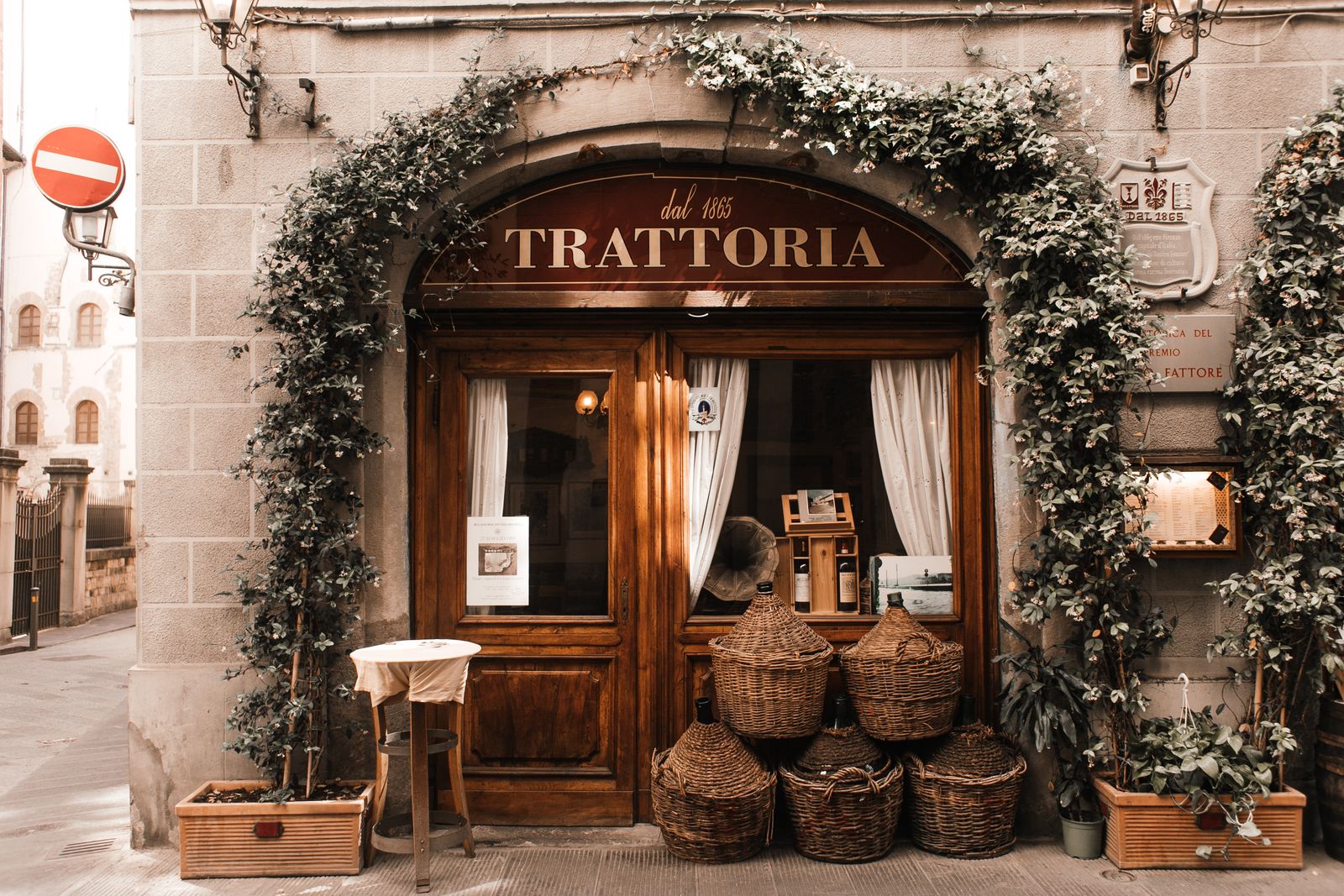 Ingresso di una trattoria italiana con porta in legno, rampicanti e damigiane impagliate
