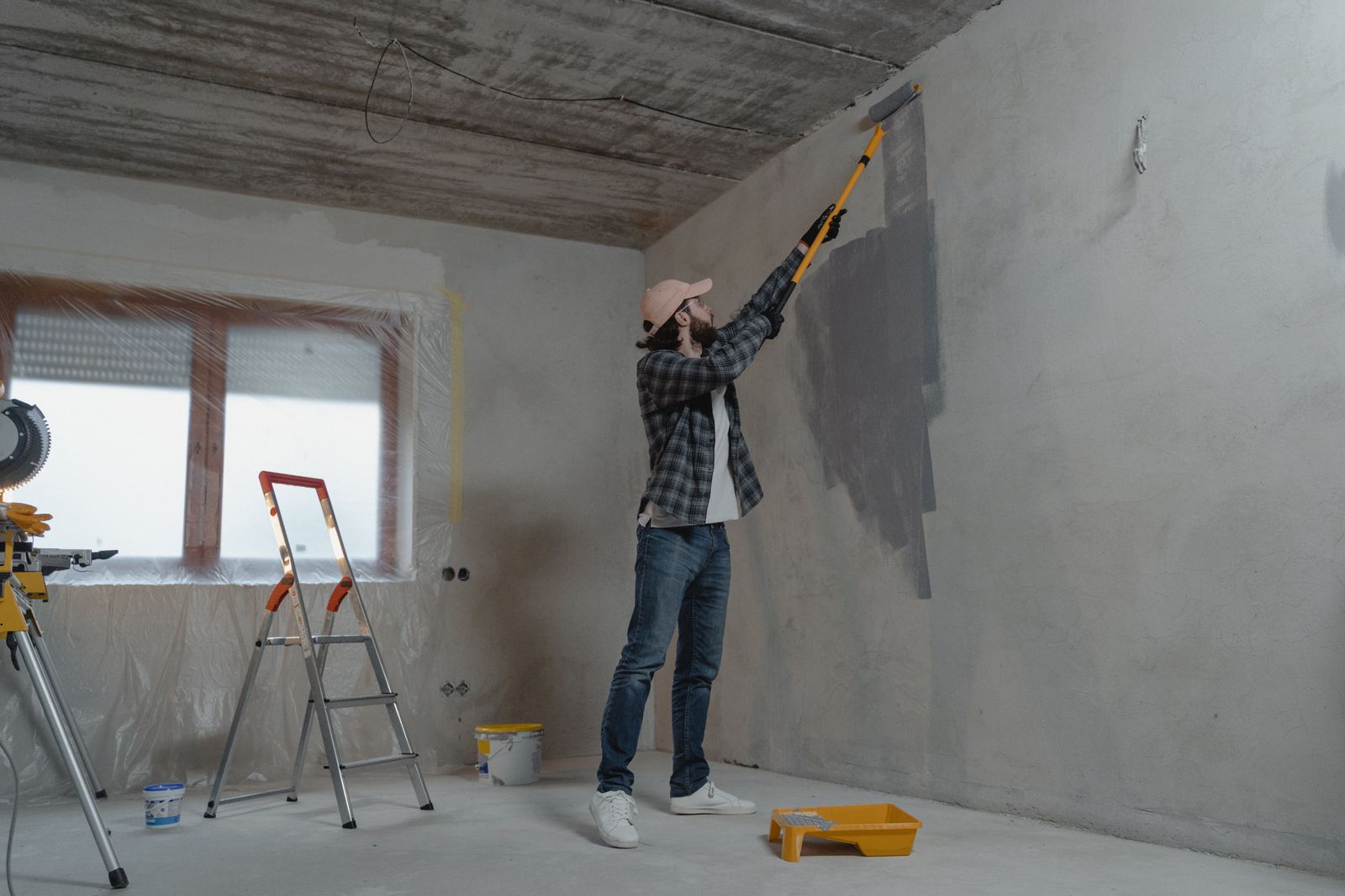 Operaio dipinge con il rullo una parete in un appartamento in ristrutturazione, con scala e attrezzi a terra