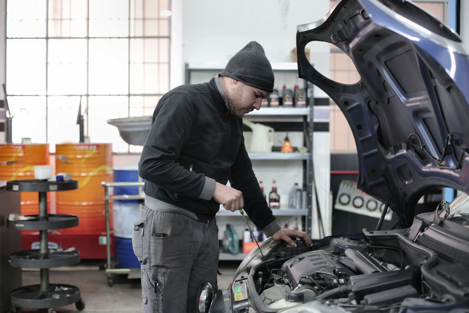 Meccanico con berretto di lana che controlla il livello dell’olio nel vano motore di un’auto con il cofano aperto in officina