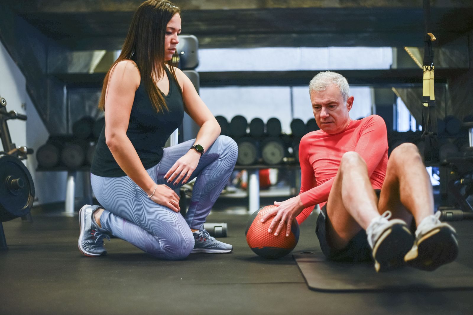 Una personal trainer inginocchiata segue un uomo di mezza età che fa un esercizio per gli addominali con la palla medica in palestra
