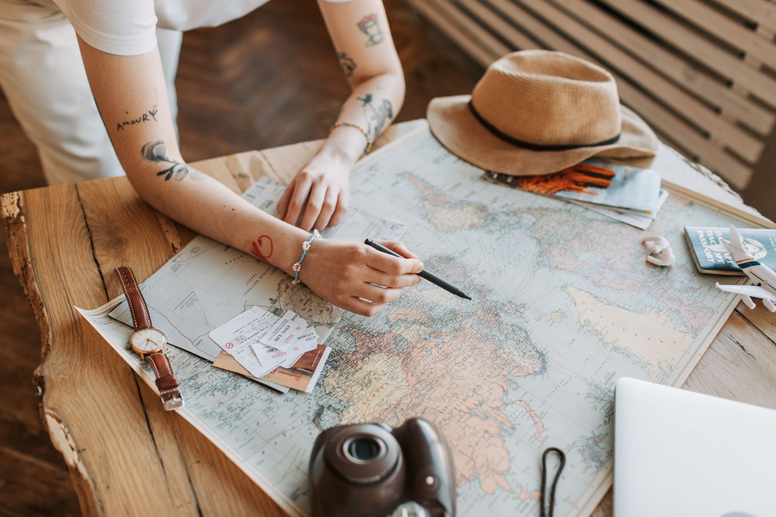 Mani che segnano un itinerario con la penna su una mappa del mondo, accanto a cappello di paglia, biglietti, orologio e macchina fotografica