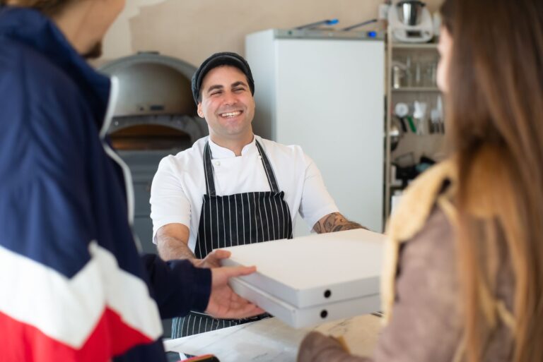 Pizzaiolo sorridente consegna due cartoni di pizza da asporto ai clienti al banco