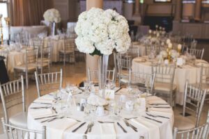 Sala ricevimenti con tavoli rotondi apparecchiati per un matrimonio, centrotavola di fiori bianchi e sedie argentate