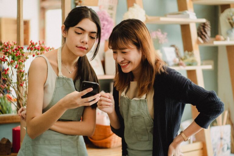 Due fioriste in grembiule leggono sorridendo un messaggio sullo smartphone all'interno del negozio